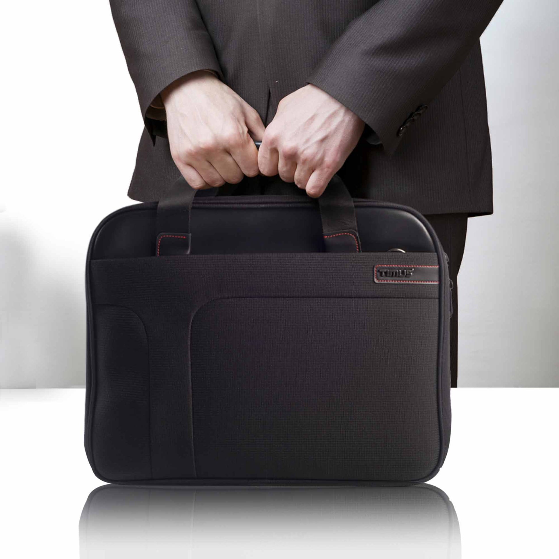 Person wearing a dark suit holding the black Timus Connect laptop carry-on bag by its dual handles, showcasing the front pocket, red stitching, and Timus logo