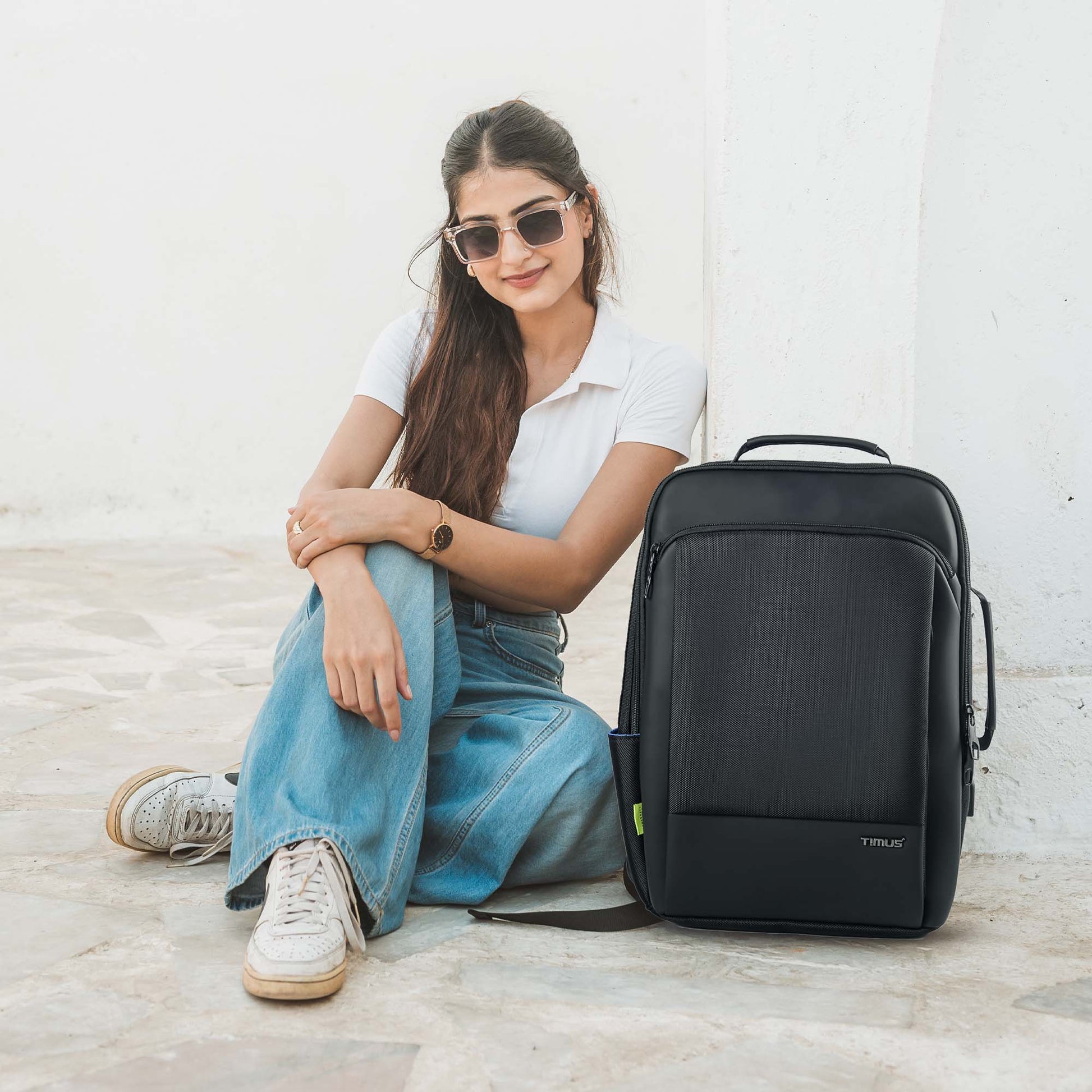 Woman sitting outdoors wearing sunglasses and white top beside the black Paris laptop backpack by Timus, showing its front view.