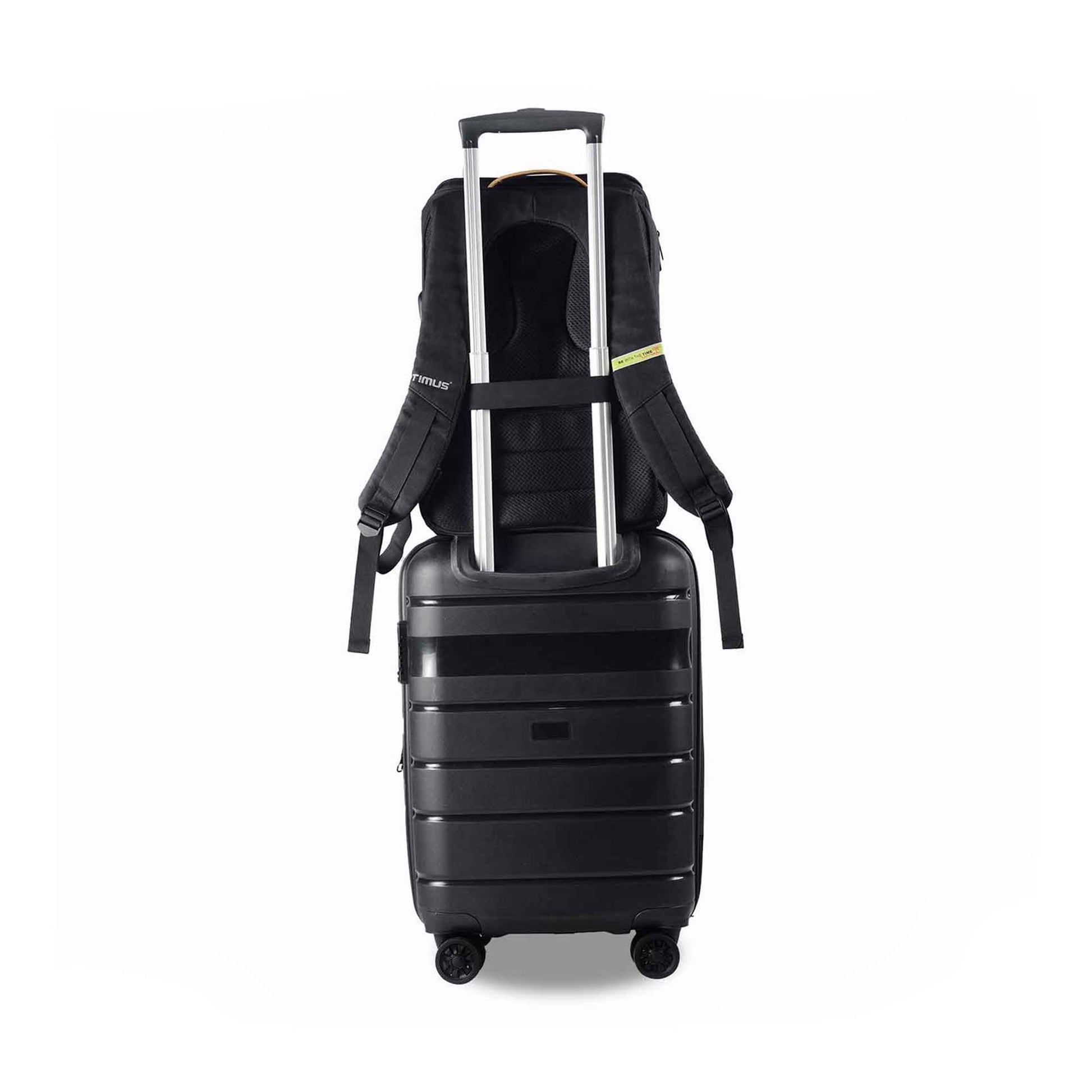 Back view of the Timus Magnet Overnighter backpack attached securely to the handle of a black rolling suitcase using its luggage strap. The suitcase has four wheels and is placed against a white background.