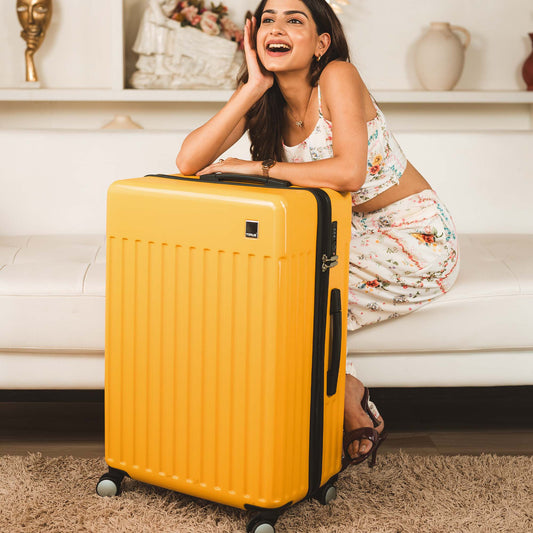 Smiling woman sitting indoors next to a Timus Neolite yellow hard shell trolley luggage with 360 degree rotation wheels and vertical textured design.